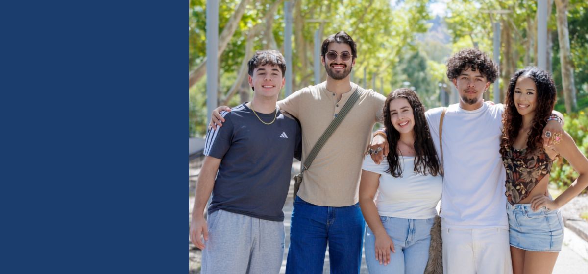 Five people pose together outdoors on a tree-lined campus walkway, smiling at th