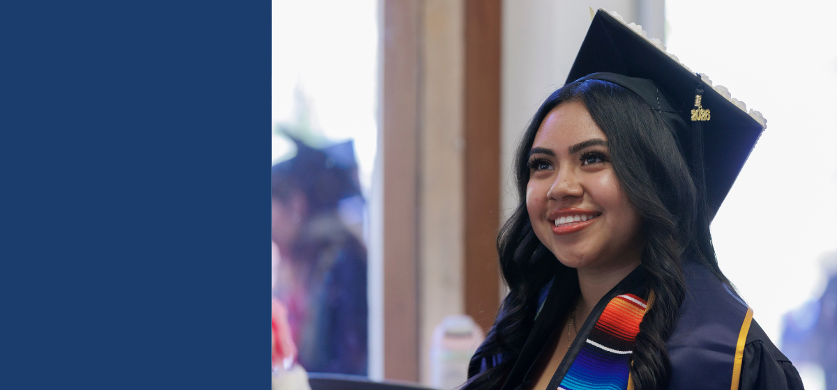 A smiling graduate wearing a black cap and gown and a colorful graduation stole 