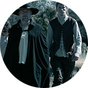 Black and white image of film with man in hat and cloak standing in a field next to a younger man in a vest.